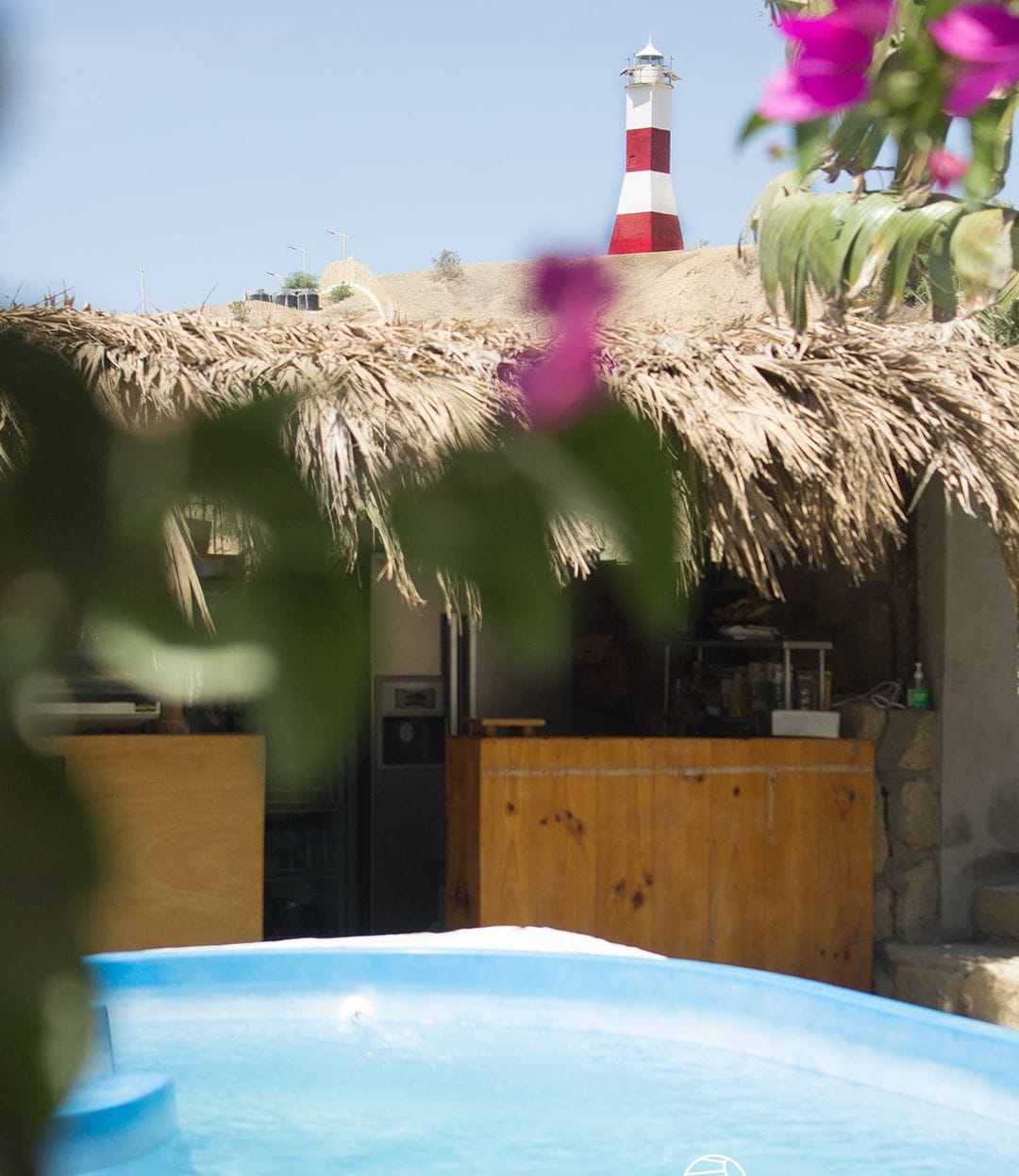 A pool area with a thatched roof structure, a lighthouse in the background, and a wooden cabin.