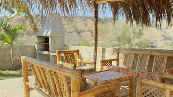 A covered outdoor seating area with bamboo furniture, a grill, and a view of a desert landscape.