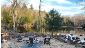 Una cabaña junto al lago con un pozo de fuego y sillas cerca del agua.