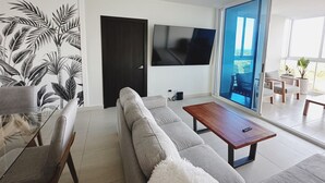 Una sala moderna con un sofá gris, una mesa de centro de madera, un televisor de pantalla plana y un ventanal con vistas al exterior.