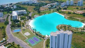 Vista aérea de una zona residencial costera con una amplia piscina turquesa, canchas de tenis y edificios altos.