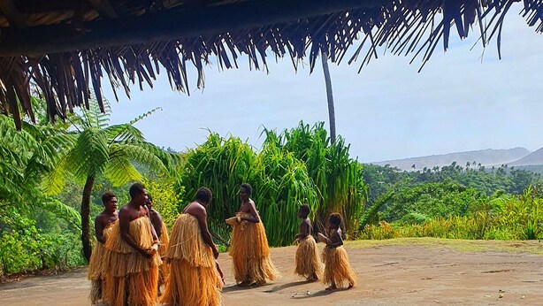 Un grupo de personas vestidas con atuendos tradicionales posan bajo una estructura de techo de paja en un entorno natural, con vegetación exuberante y colinas al fondo.