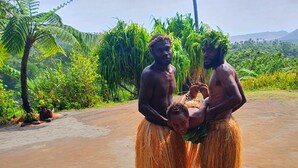 Tres personas vestidas con atuendos tradicionales sobre un sendero de tierra, rodeadas de vegetación exuberante y una montaña al fondo.