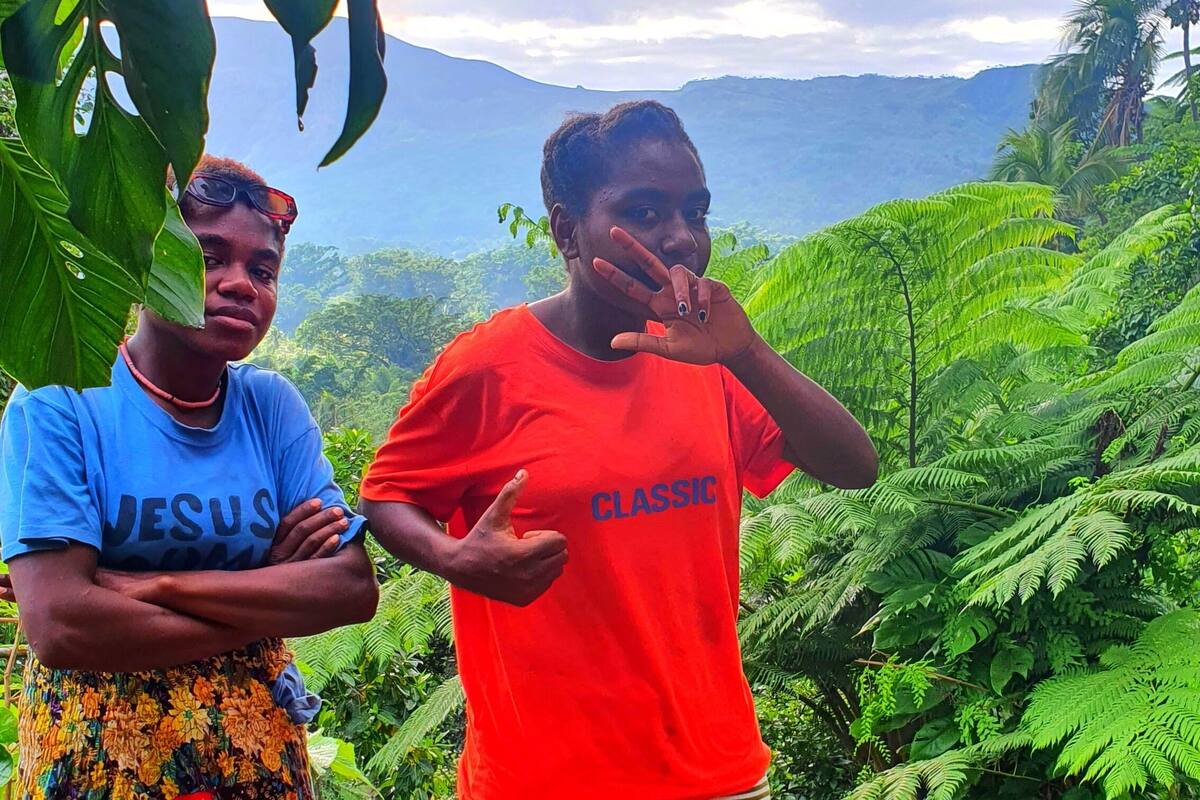 Two individuals standing outdoors with lush greenery and mountains in the background.