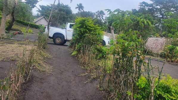 Un chemin de terre mène à un pick-up blanc garé dans une zone rurale, entourée d’une végétation dense, avec une construction à toit de chaume en arrière-plan.