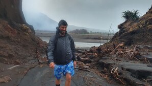 Iemand loopt over een rotsachtig pad, met een rivier en bergen op de achtergrond.