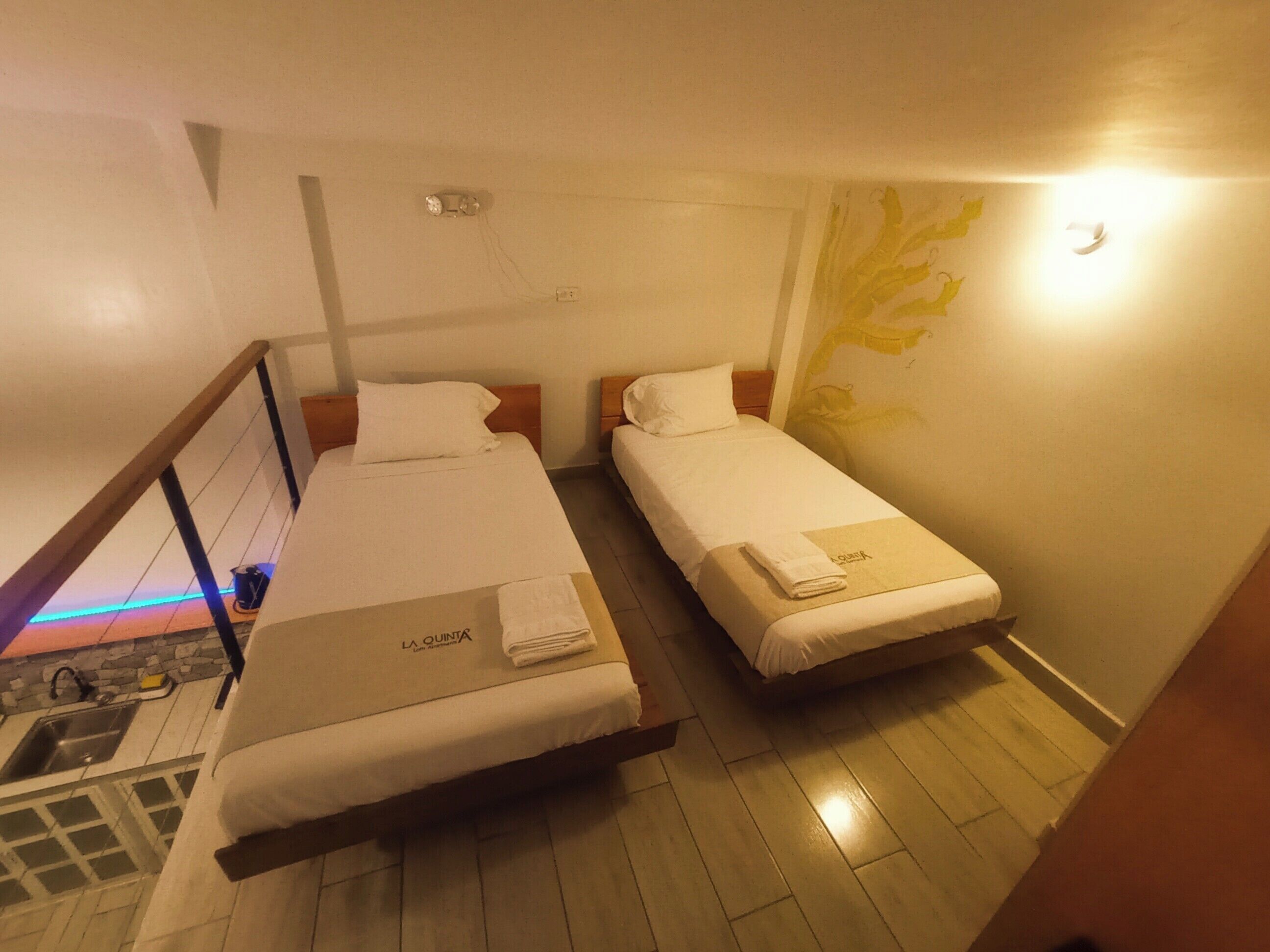 A hotel room with two single beds, a wooden floor, a small bathroom with a sink, and a wall-mounted light fixture.