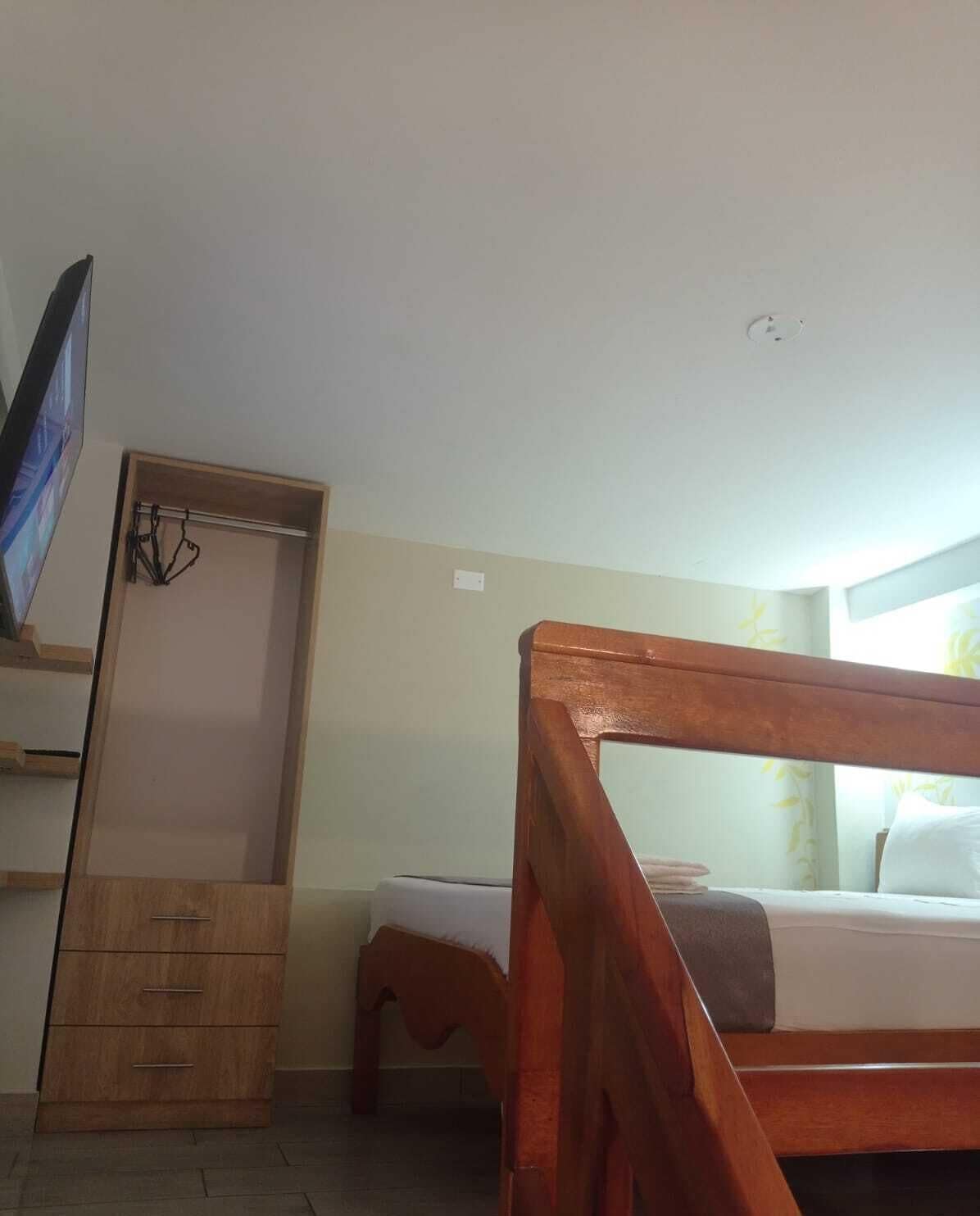 A bedroom with a wooden bed frame, a TV mounted on the wall, a wooden wardrobe, and a small bedside table.