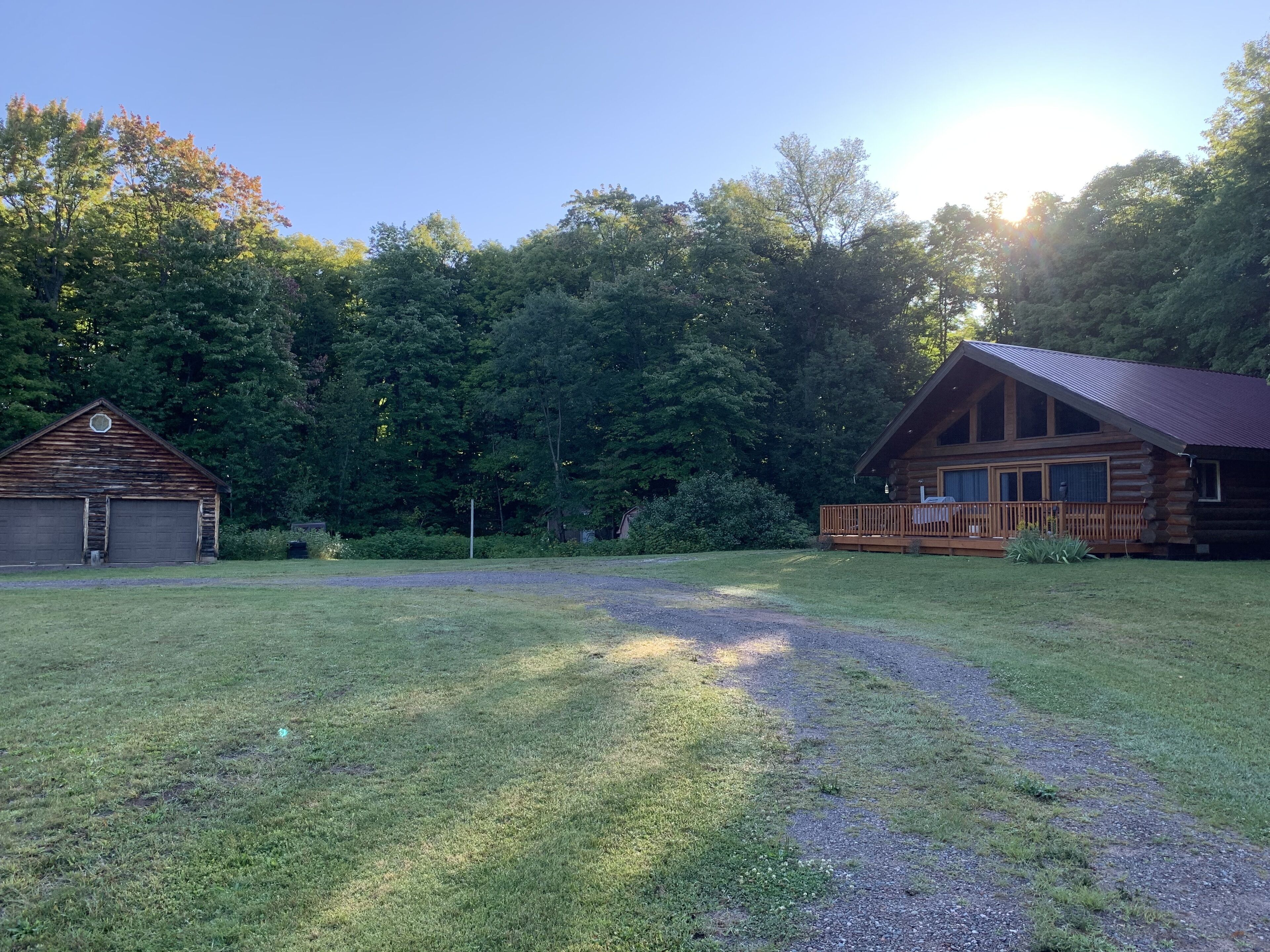 A rustic cabin with a porch and a garage, surrounded by a grassy yard and dense trees.