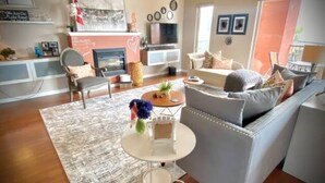 A living room with a grey sofa, a coffee table, and a TV. There is a fireplace, a bookshelf, and a window with curtains. - Osoyoos Lakeside 2 BDRM Condo! (Osoyoos)