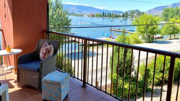 A balcony with wicker furniture, a view of a lake with boats, and a mountainous landscape in the background. - Osoyoos Lakeside 2 BDRM Condo! (Osoyoos)