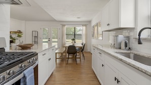 Una cocina moderna con armarios blancos, encimera de madera, cocina a gas y comedor con mesa y sillas.