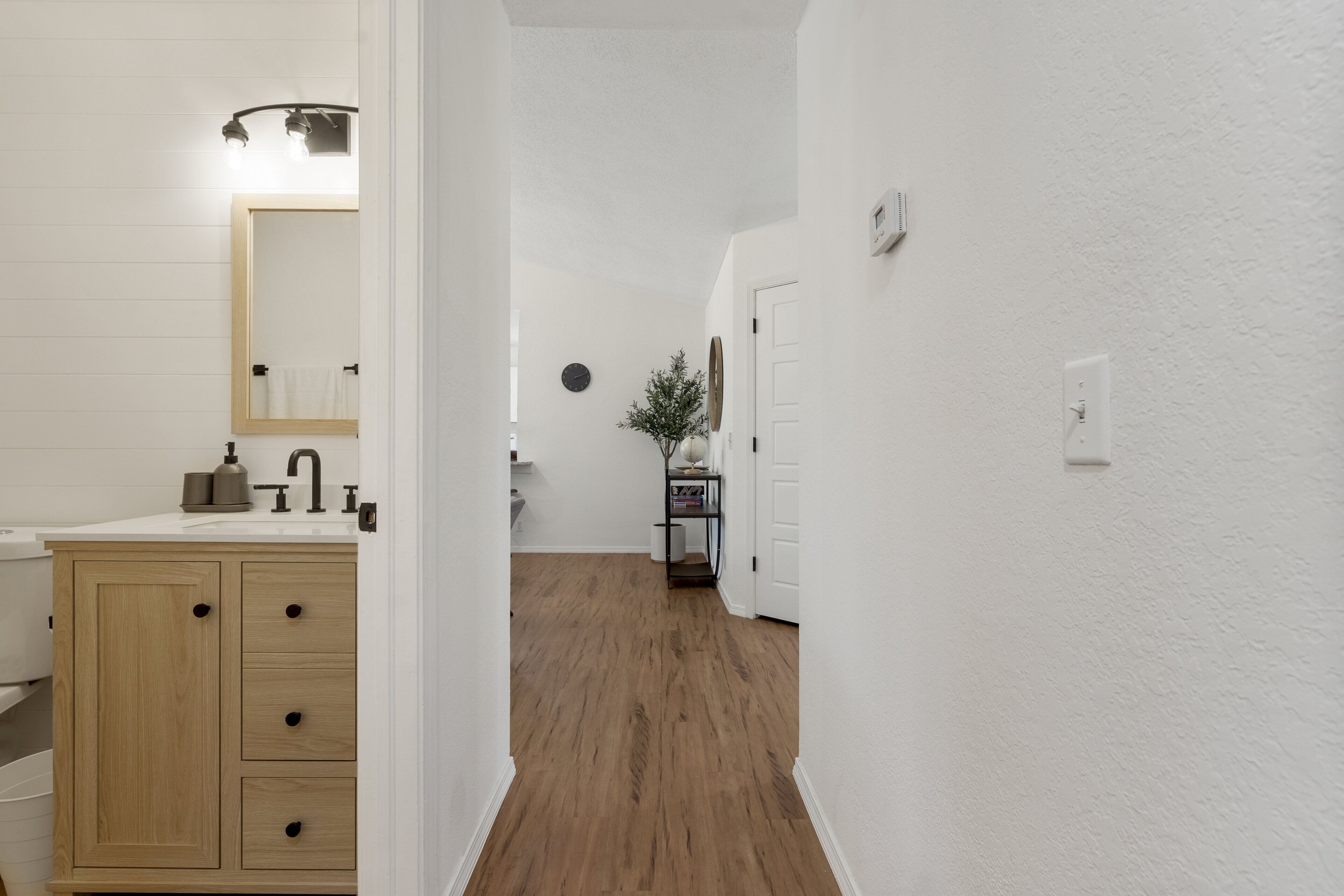 A hallway with a wooden floor, white walls, a wooden cabinet with drawers, a mirror, and a small table with a plant.
