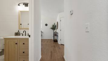 A hallway with a wooden floor, white walls, a wooden cabinet with drawers, a mirror, and a small table with a plant.