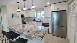 A modern kitchen with white cabinets, a granite countertop, a stainless steel refrigerator, and pendant lighting. - CP 326 Ocean View Condo- Welcome to Paradise (Fort Pierce)