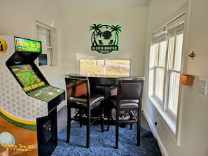 Ein Zimmer mit einem 3D-Golf-Arcade-Spiel, einem Tisch mit vier Stühlen und dem an der Wand befestigten Logo von Hamm House.