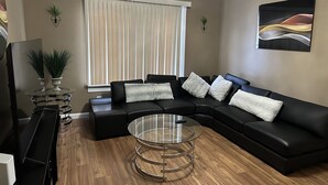 A modern living room with a sectional sofa, a glass coffee table, and a large window with blinds.