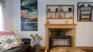 A living room with a fireplace, a couch, a round wooden table, and artwork on the walls. - Cosy Character Cottage in the City | Walk, Ride & Unwind (Rotorua)