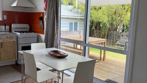 A modern kitchen with a gas stove, oven, and a white cabinet. There is a dining table with chairs and a large window overlooking a backyard with a wooden deck and a house. - Cosy Character Cottage in the City | Walk, Ride & Unwind (Rotorua)