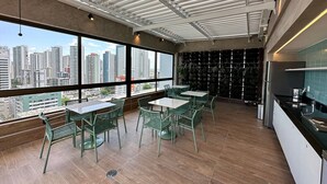 A modern dining area with tables and chairs, large windows offering a city view, and a kitchen area with a sink and countertop. - APARTAMENTO ALTO LUXO (Recife)