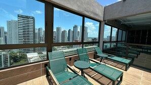 A rooftop terrace with green chairs and tables, large windows offering a city view. - APARTAMENTO ALTO LUXO (Recife)