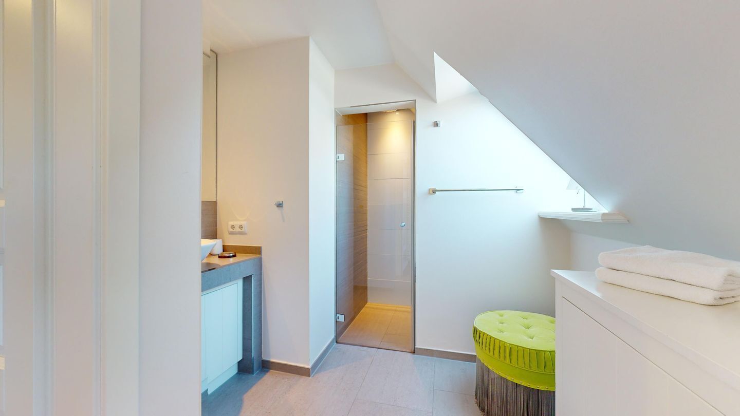 A narrow hallway leading to a bathroom with a glass shower door and a small green stool.