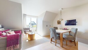 Un salon moderne avec un canapé, une table basse et un coin repas avec des chaises. Une grande fenêtre offre une vue sur l’extérieur.