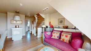 Una sala moderna con una escalera de madera, un sofá con cojines estampados y un comedor con un armario blanco y una lámpara colgante.