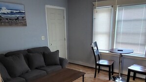 Un salon avec un canapé gris, une table basse en bois, un ventilateur de plafond et une fenêtre avec des stores.