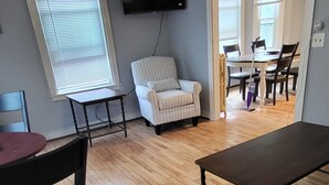 Un salon doté d’un plancher en bois, d’un ventilateur de plafond, d’un téléviseur fixé au mur et d’un coin repas avec une table et des chaises.