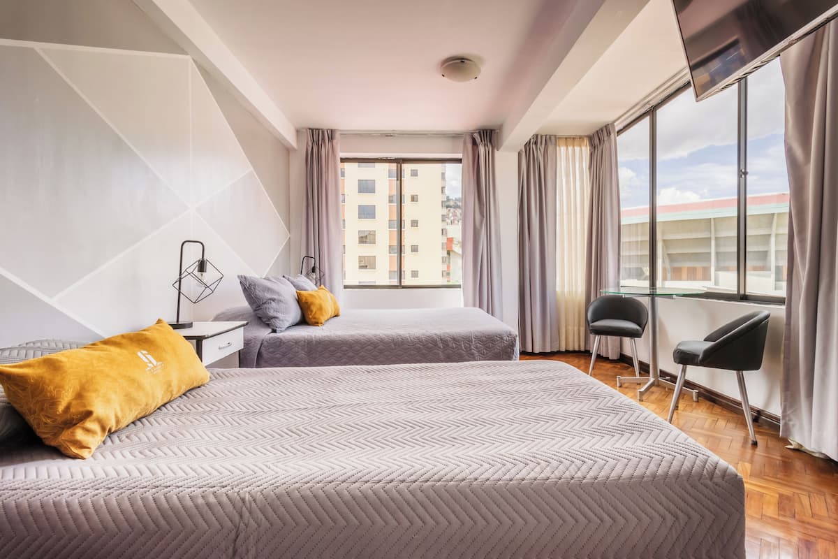 A hotel room with two beds, a desk, a chair, a TV, and a large window with curtains.