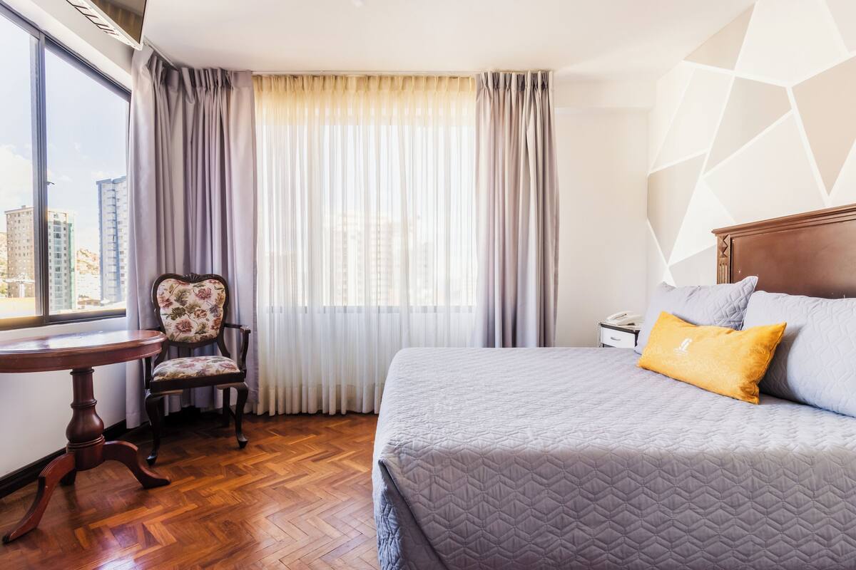 A hotel room with a bed, a chair, a table, and a window with curtains.