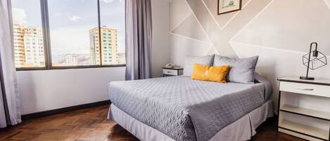 Una habitación de hotel moderna con una cama grande, una mesita de noche, un escritorio y una ventana con vistas a la ciudad.