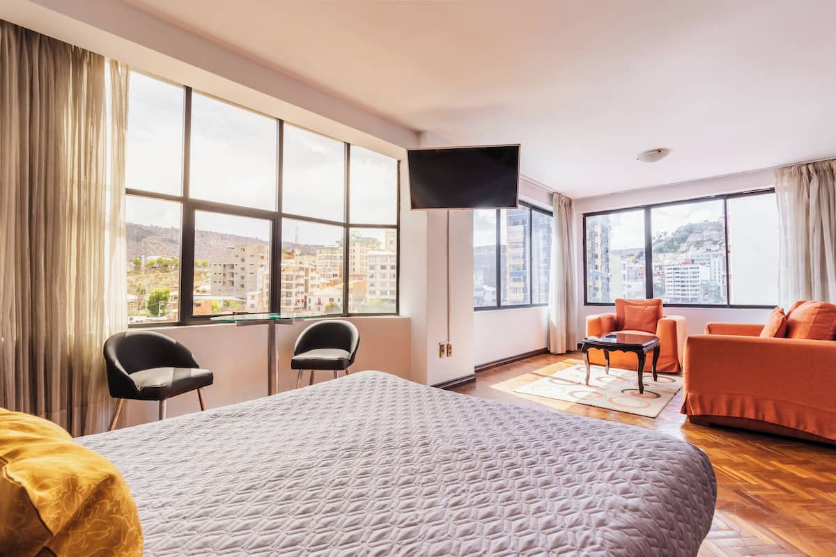 A hotel room with a large bed, two chairs, a sofa, a flat-screen TV, and large windows offering city views.