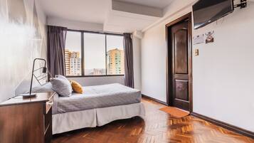 A bedroom with a bed, a desk, a chair, a TV mounted on the wall, and a view of buildings outside.