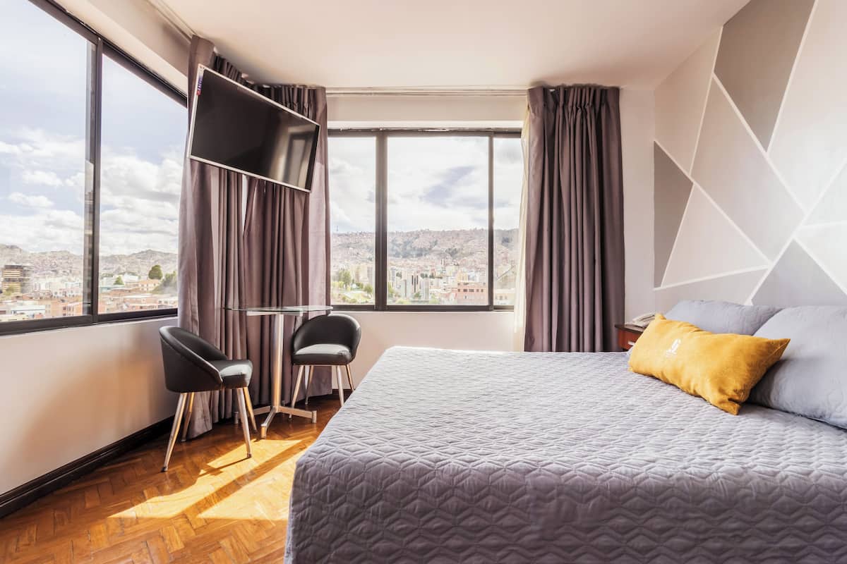 A hotel room with a bed, a desk, a chair, and a large window offering a city view.