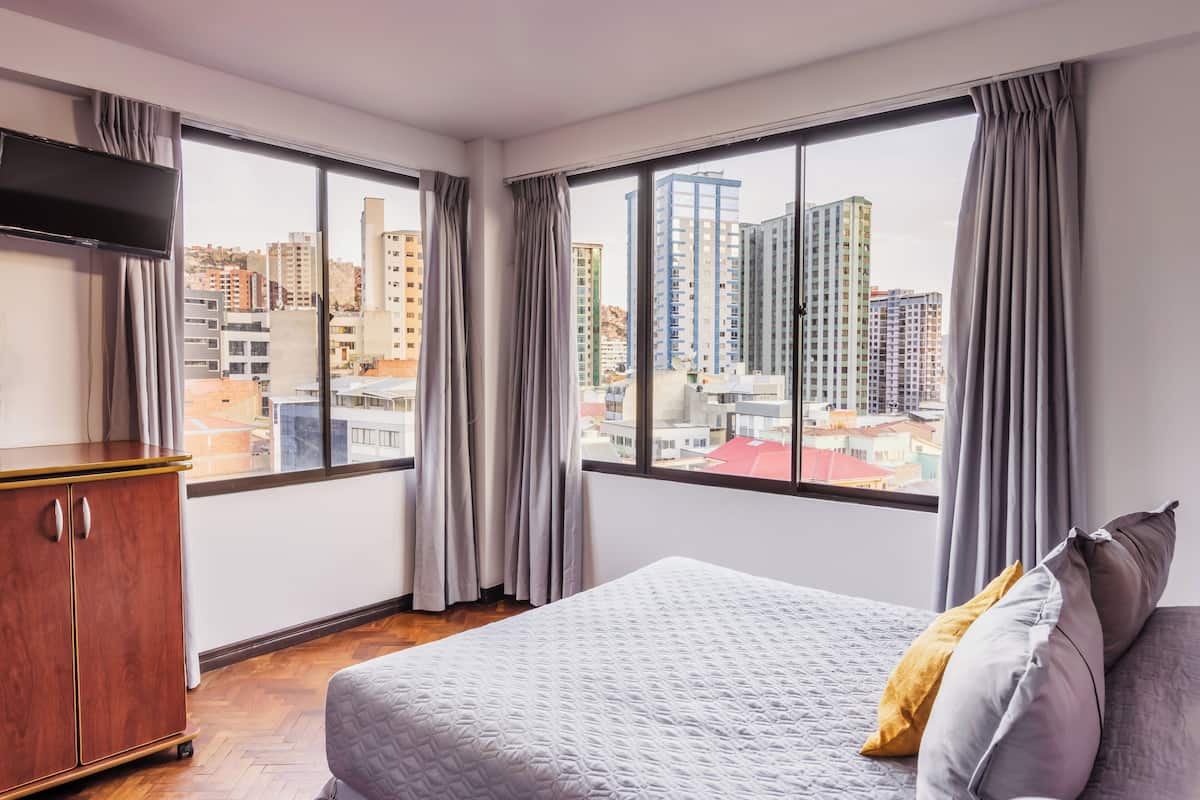 A hotel room with a bed, a TV, a wooden cabinet, and large windows offering a city view.