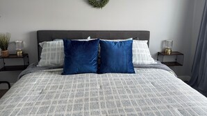Un dormitorio con cabecera gris, colcha estampada en gris y blanco, dos almohadas azules, una guirnalda verde en la pared y dos mesitas de noche con lámparas y una planta en maceta.
