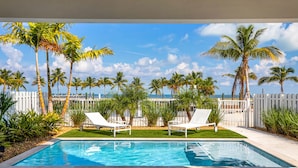 Piscina com duas espreguiçadeiras brancas, cercada por palmeiras e um muro branco, com vista para o oceano.