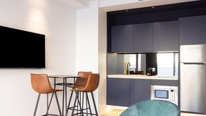 Eine moderne Küche mit dunklen Schränken, einem Edelstahl-Kühlschrank und einer eingebauten Mikrowelle. Ein runder Tisch mit zwei Stühlen und ein petrolfarbener Ohrensessel sind vorhanden.