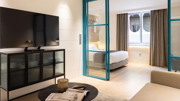 A modern hotel room with a large flat-screen TV, a glass cabinet, a round coffee table with magazines, a bed with a view, and a teal-framed sliding door. - Chayofa Suites - Deluxe (Santa Cruz de Tenerife)