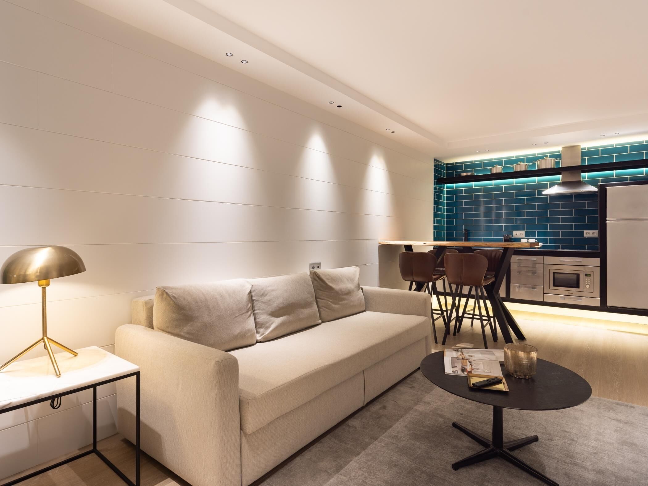 A modern living room with a sofa, a coffee table, and a lamp. There is a kitchen area with blue tiles and a refrigerator.