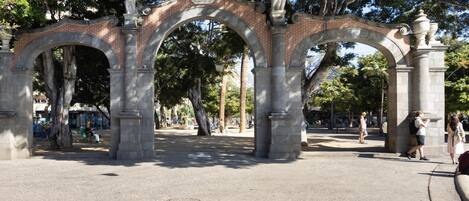Un arco de piedra con columnas, rodeado de árboles y una zona pavimentada.