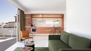 Uma sala de estar moderna com um sofá verde, uma cadeira marrom e uma mesa de centro de madeira. Uma cozinha com mesa e cadeiras é visível ao fundo.