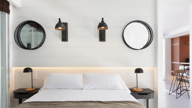 Quarto moderno com uma cama grande, duas mesas de cabeceira com luminárias e três espelhos fixados na parede.