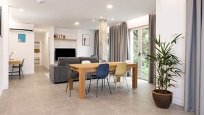 A modern living room with a wooden dining table, chairs, a sofa, and a television. There is a potted plant and a large window with curtains. - ★ New · Guimera Family Suites - A (Santa Cruz de Tenerife)