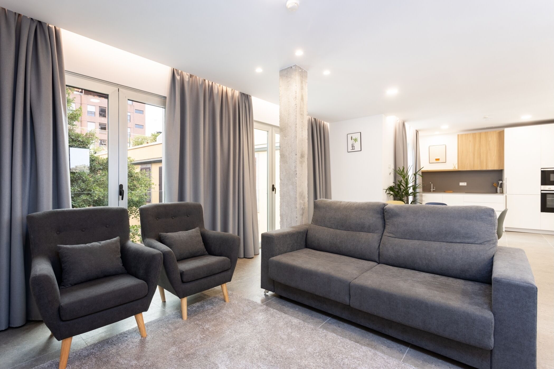 Un salon moderne avec un canapé gris et deux fauteuils, un pilier en béton et une vue sur l’extérieur grâce à de grandes fenêtres.