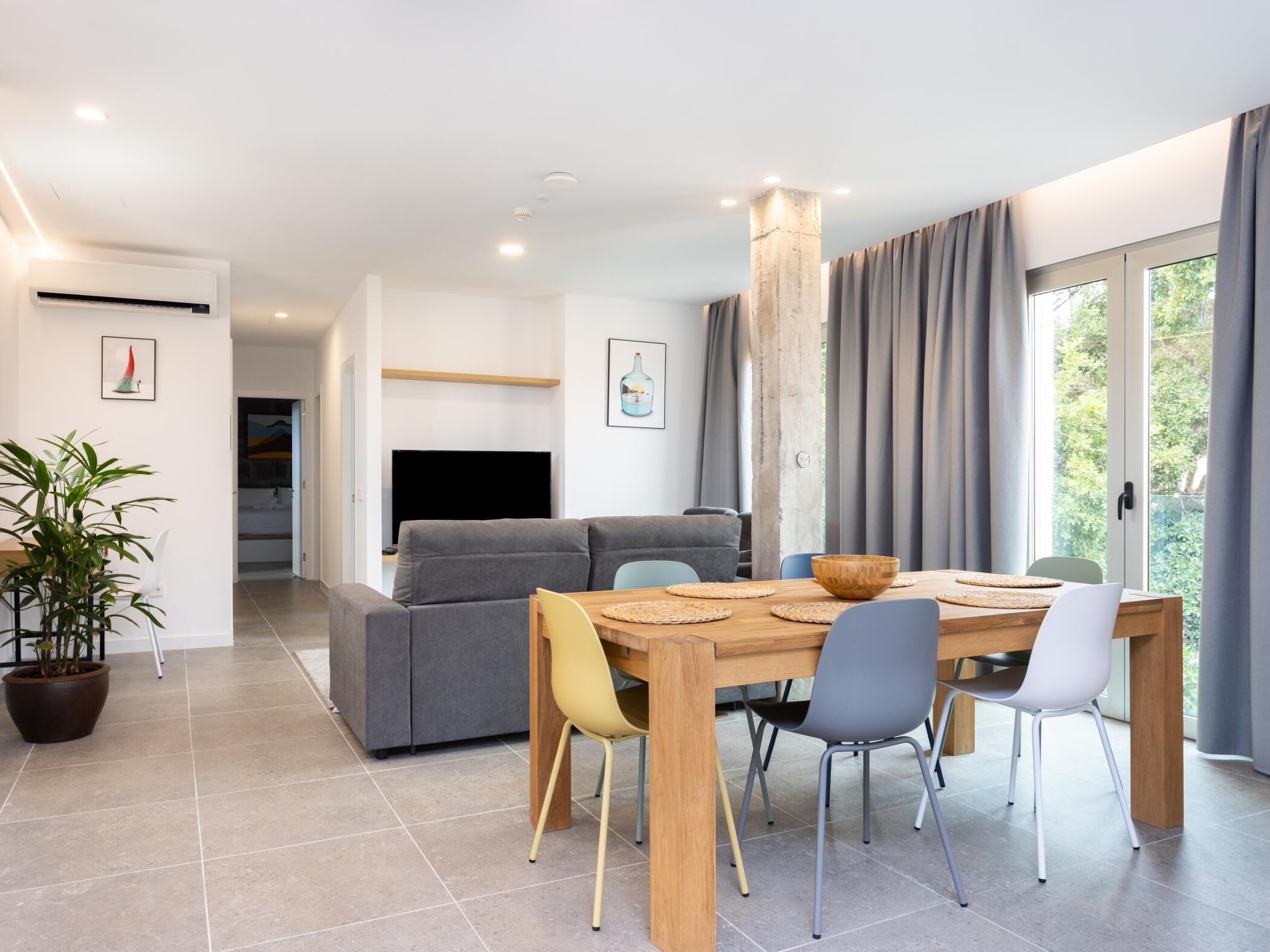 A modern living room with a grey sofa, a wooden dining table with chairs, a TV, and a large window with curtains.