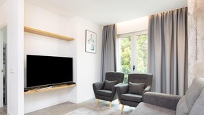 Ein modernes Wohnzimmer mit einem an einer Holzablage montierten Flachbildfernseher, zwei grauen Sesseln, einem grauen Sofa und einem großen Fenster mit Vorhängen.