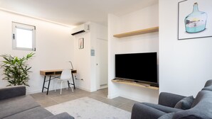Ein modernes Wohnzimmer mit Flachbildfernseher, grauen Sofas, einem Schreibtisch mit Stuhl, einer Topfpflanze und einem Bild an der Wand.